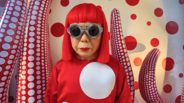 Yayoi Kusama com peruca vermelha, blusa vermelha com um círculo gigante e óculos, em frente a uma obra de arte vermelha e branca com círculos, que lembra um tentáculo