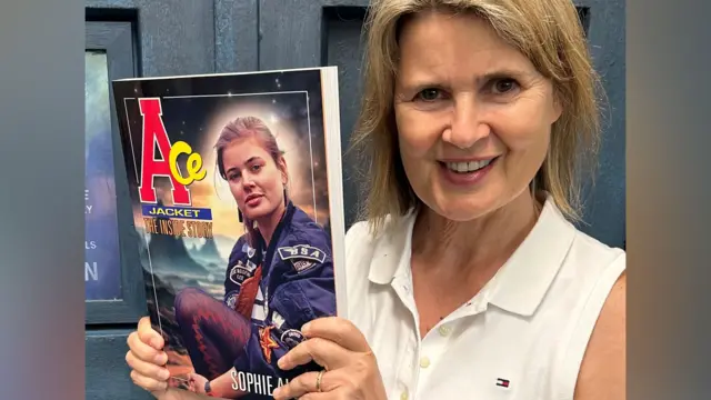 Sophie Aldred is holding a copy of her book in front of the blue doors of a police box. She is smiling and wearing a white top. The book features a picture of Sophie in her Ace costume as a younger woman.