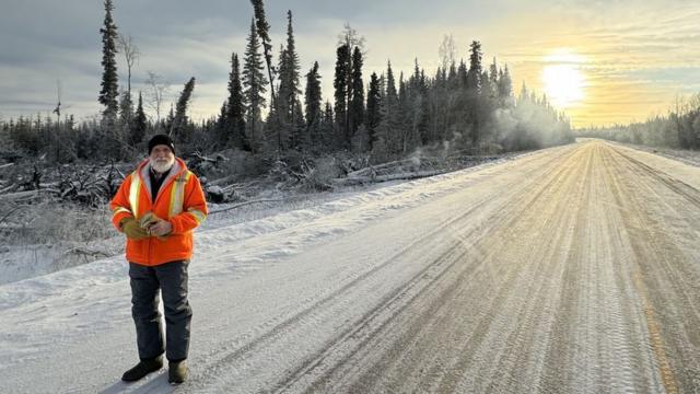 Foto de senhor branco idoso com roupas de frios em uma estrada com uma paisagem coberta com neve, mas com fumaça ao fundo 