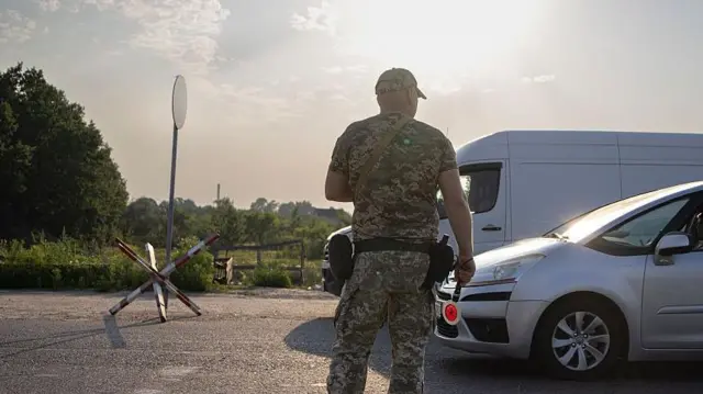Чоловік у військовому камуфляжі стоїть спиною до камери посеред дороги біля імпровізованого блокпоста. Праворуч зупинений легковий автомобіль, у вікні якого видно водія. Позаду військовослужбовця – білий фургон. На узбіччі – дерев’яний шлагбаум, а вдалині – зелені дерева та відкрита місцевість.