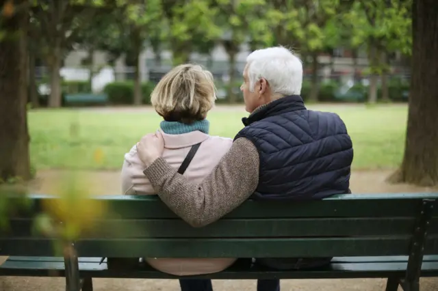 Una pareja mayor está sentada en una banca en un parque de espaldas a la cámara.