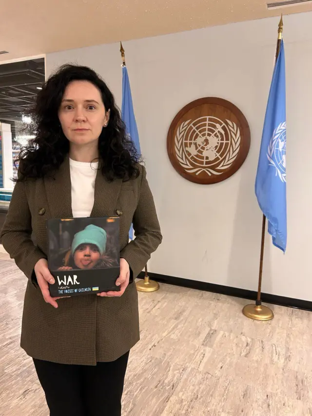 Елена Розвадовская в коричневом пиджаке с книгой в руках. На обложке книги девочка показывает язык