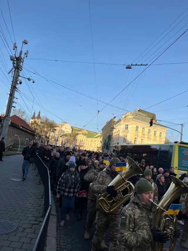 Десятки людей супроводжують похоронну процесію на кладовище.