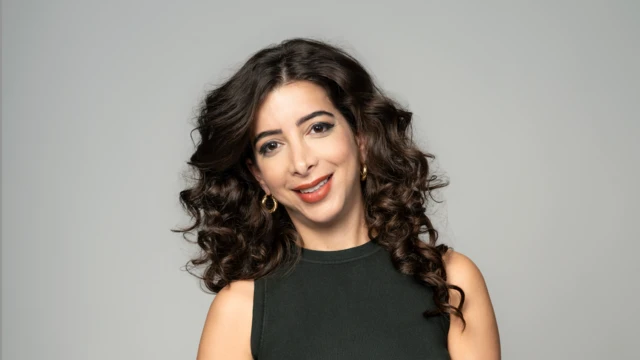 A woman with black curly hair, wearing a black tank top and gold hoop earrings.