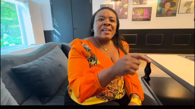 Lydia Mutyebele Ngoi mw'ishati y'ibara ry'umucungwe irimwo n'amabara y'igitenge, yicaye muri diva muri imwe mu mahoteri ya Bruxelles mu 2025