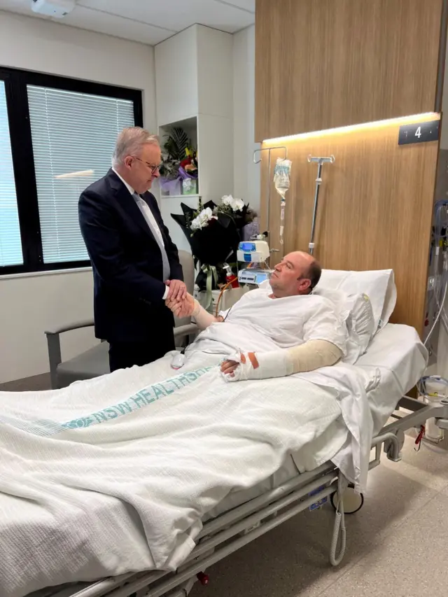 病室のベッドに横たわる男性の横に、スーツ姿の男性が立ち、負傷した男性の右手を両手で持っている。病床の男性は左腕をギプスで固定している