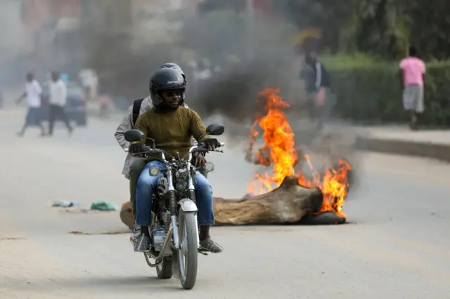 Два человека едут на мотоцикле на фоне чего-то горящего и дымящегося на дороге