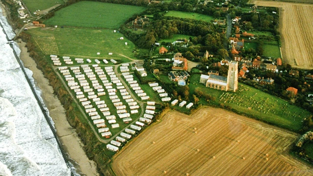 Una imagen aérea de una costa, una playa y un pueblo, que muestra un gran campo y un parque de caravanas directamente adyacente a los acantilados y más allá de eso una iglesia, un cementerio y viviendas.