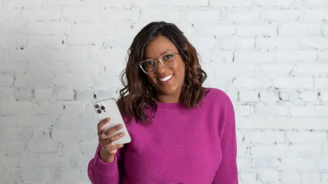 Andréa Jones sorrindo, segurando o celular e vestindo uma blusa roxa.