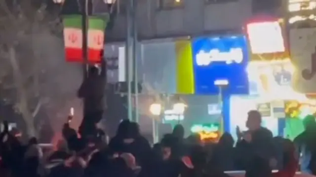 Un homme grimpe sur un poteau sur lequel est hissé le drapeau iranien après la tombée de la nuit. De nombreux manifestants sont rassemblés autour de lui. Cette image a été publiée sur les réseaux sociaux le 8 janvier 2026.