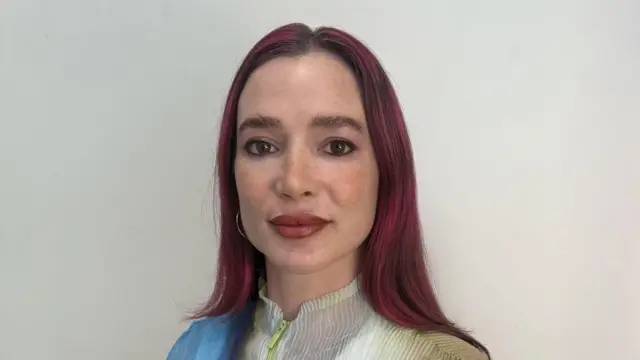 Uma mulher de cabelos longos vermelho-escuros olha para a câmera.