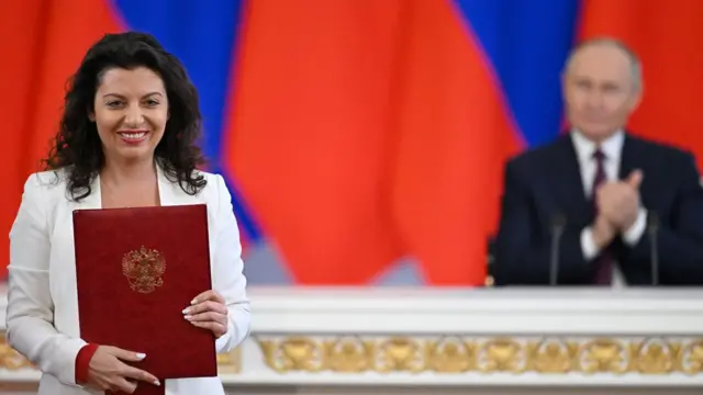 Головна редакторка RT Маргарита Симоньян з бордовою папкою з гербом РФ, на задньому плані президент Росії Володимир Путін