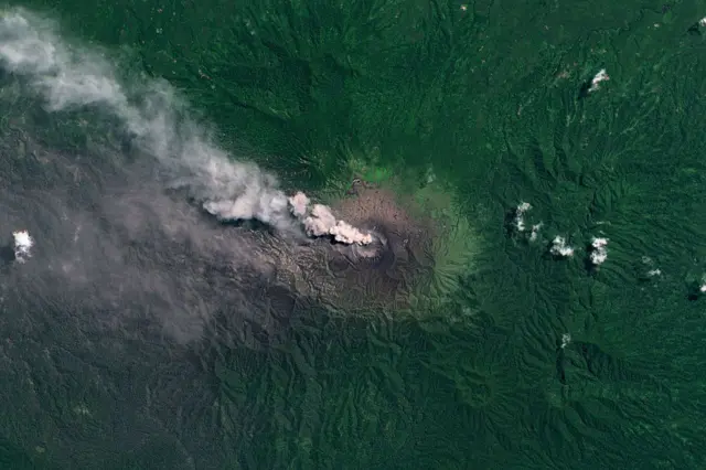 Satellite image of Mount Dukono showing plumes of smoke coming out of the volcano's crater, surrounded by dense green vegetation