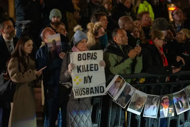 Protestocu bir grup insan ellerinde fotoğraflar ve dövizler tutuyor. Bir kadın "Boris kocamı öldürdü" diyerek eski başbakan Boris Johnson'ı protesto ediyor.