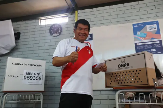 Un hombre vestido con la camiseta de la selección peruana de fútbol posa sonriente y con un pulgar alzado en un centro de votación en Lima.
