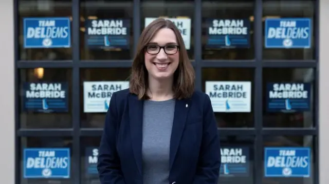 Sarah Mcbride posa para una fotografía frente a varios letreros que llevan su nombre. Viste con una chaqueta azul, camisa gris, anteojos azules y tiene el cabello hasta las mejillas. 