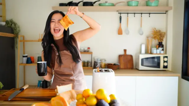 Молода жінка в фартуху готує сніданок і насолоджується чашкою кави вранці у своїй кухні - стокова фотографія