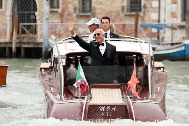 Jeff Bezos wear dark glasses and tuxedo as e wave from di back of a small motor boat for Venice on 27 June 2025.