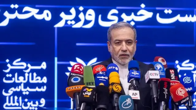 Седой мужчина с бородкой и усами перед множеством микрофонов