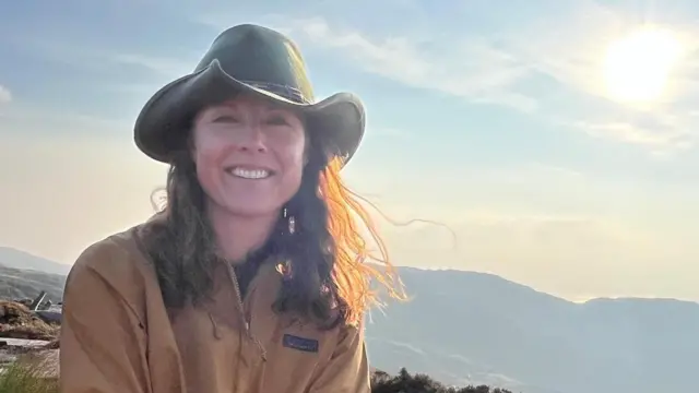 니나 라이언이 모자를 쓰고 카메라를 향해 미소 짓고 있으며, 뒤로는 산의 능선이 보인다