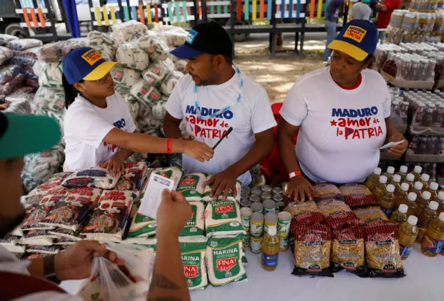 Três pessoas atrás de mesa com produtos básicos; elas vestem camisetas brancas de apoio a Maduro