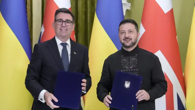 Бернем (ліворуч) і Зеленський (праворуч) у формальному одязі стоять перед прапорами України та Великої Британії. Кожен тримає синю папку з документами.