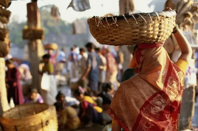 ਇਕੱਠ ਵਿੱਚ ਇੱਕ ਔਰਤ ਸਿਰ ਉੱਤੇ ਟੋਕਰਾ ਚੁੱਕੀ ਜਾ ਰਹੀ ਹੈ, ਮੂੰਹ ਦੂਜੇ ਪਾਸੇ ਹੈ, ਧੁੰਦਲੇ ਪਿਛੋਕੜ ਵਿੱਚ ਬਜ਼ਾਰ ਵਰਗਾ ਦ੍ਰਿਸ਼ ਹੈ
