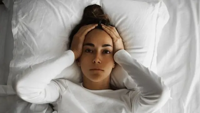 امرأة تستلقي على سرير أبيض وتضع يديها على رأسها ويبدو عليها عدم الارتياح