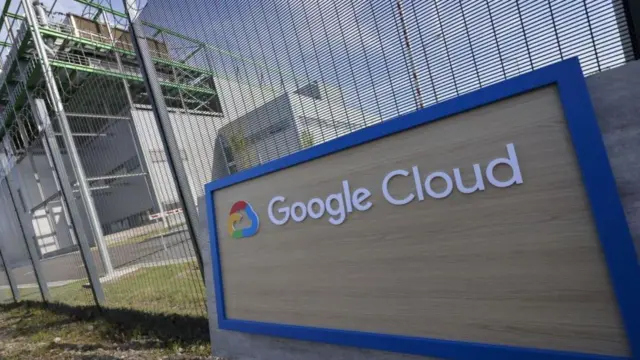 لوحة تحمل شعار غوغل كلاود باللغة الإنجليزية، لون الخط أبيض على لوحة بلون قمحي خشبي وإطار أزرق غامق، بجانب الشعار رمز سحابة ملون بالأحمر والأزرق والأخضر. اللوحة أمام سياج معدني، وفي الخلفية يقع مركز بيانات. الصورة التقطت نهاراً.
