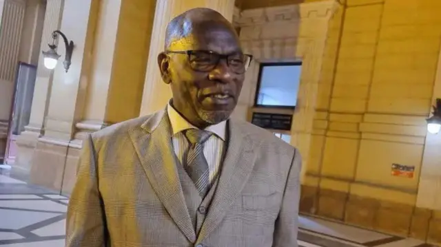 Séraphin Twahirwa, ahagaze amwenyura, yambaye ikoti na 'cravate', inyuma ku ruhande rumwe hari itara ryaka, no ku rundi hari itara ryaka