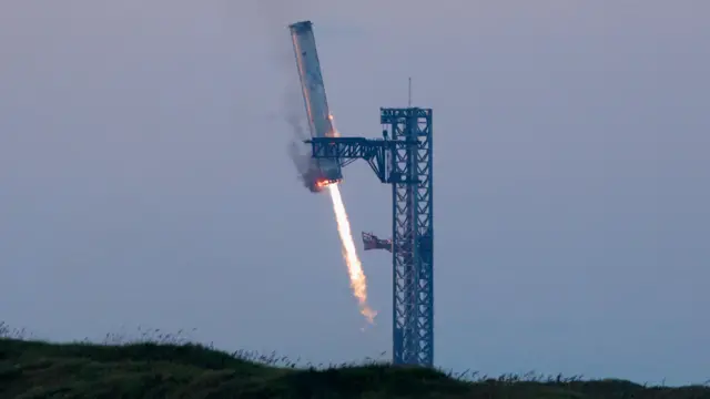 Starship's booster at an angle as it descends to the launch pad at an angle with a long thin jet of burning fuel below it