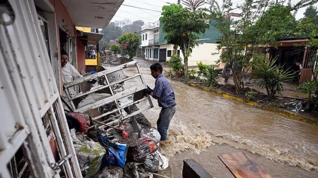 Dos hombres intentan despejar escombros de una casa, mientras uno está parado con el agua hasta las rodillas en una calle inundada