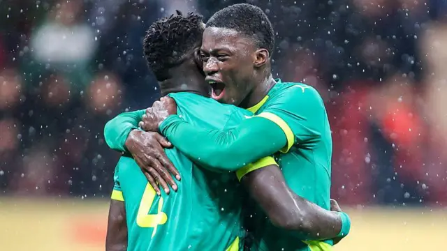 On voit Mamadou Sarr, en buste, enlaçant son coéquipier sénégalais Abdoulaye Seck après la finale de la CAN 2025. Sarr, à droite, les bras levés autour du dos de Seck, semble crier, la bouche ouverte et les yeux fermés, tandis que sa tête cache le visage de Seck. Tous deux portent le maillot vert du Sénégal et de grosses gouttes de pluie tombent autour d'eux.