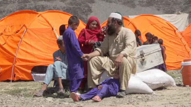 مردی با ریش با یک پیراهن و شلوار به رنگ بژ و یک کلاه سفید روی چند گونی در فضای باز نشسته است. او دست کودکی را گرفته که پیراهن و شلوار بنفش به تن دارد. دختری با روسری قرمز بین آنها ایستاده است. یک کودک دیگر با رنگ بنفش روی زمین در کنار پای مرد دراز کشیده است. چادرهای نارنجی و یک صندوق پشت آنها قرار دارد