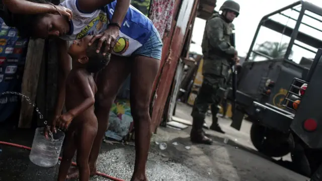 Mãe dá banho no filho na rua, pertojogo da roleta que multiplicaum militar armado