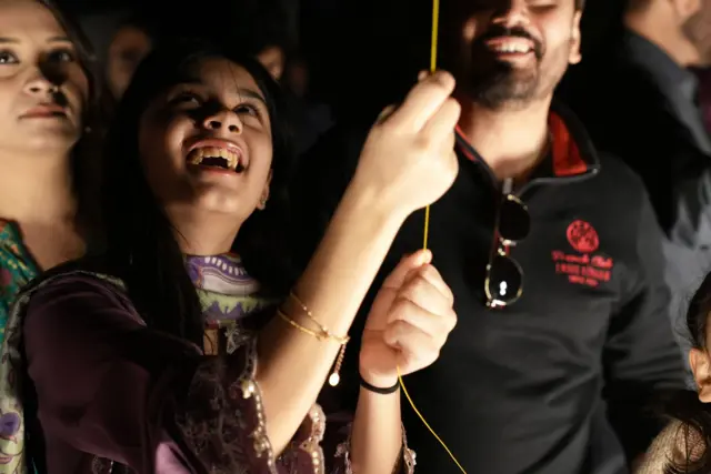 A young girl in a purple dress is holding yellow coloured kite string, smiling and looking upwards at her kite in the sky. A man overlooking her is also smiling in the direction of her kite flying in the sky.