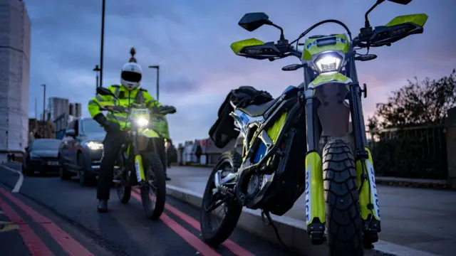 На фото в сумерках на переднем плане скутер в ярко зелено-голубых цветах полиции. На заднем плане сотрудник полиции в шлеме и ярко-зеленой форменной куртке сидит за рулем такого же скутера 