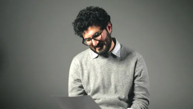 Armen Aramjan sedi u studiju, na stolici i gleda u dokumenta koja drži u desnoj ruci, ima tamnu kobvrdžavu kosu, naočale, svetlo sivi džemper i belu košulju ispod