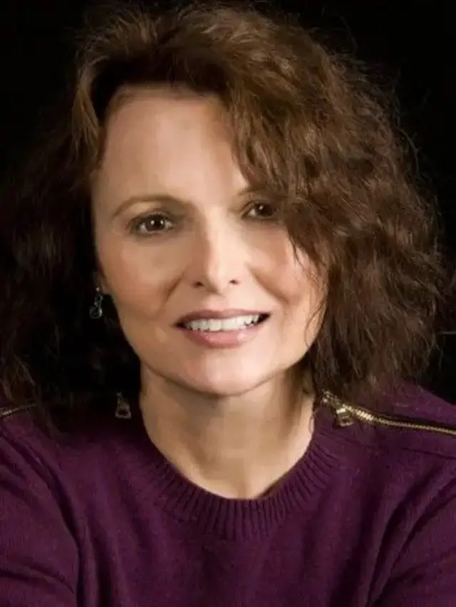 Gail é uma mulher branca, com cabelos castanhos e curtos. Ela usa um suéter roxo.