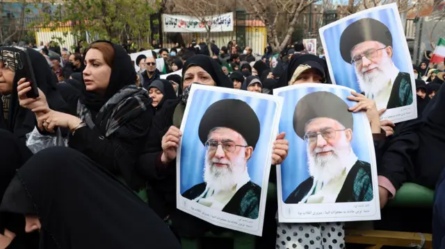 Woman dressed in black robes gather in an outdoor public area on 14 January 2026. Three of them are holding posters of Khamenei.