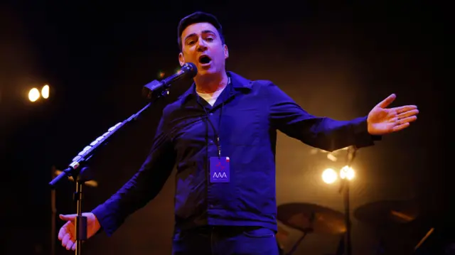 Andy Burnham no palco, com os braços abertos. Uma bateria aparece ao fundo, com luzes de palco iluminando-o. Ele está vestindo uma camiseta branca e uma camisa roxa de manga comprida com botões, em 13 de março de 2023.