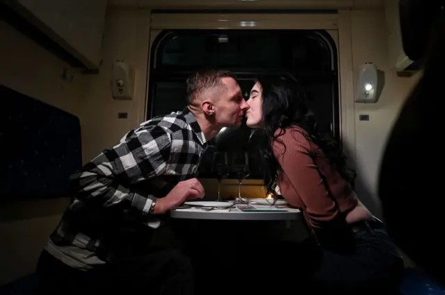 Um casal, dentro de um trem, sentado de frente para o outro, se beijando. Há taças de vinho em cima da mesa