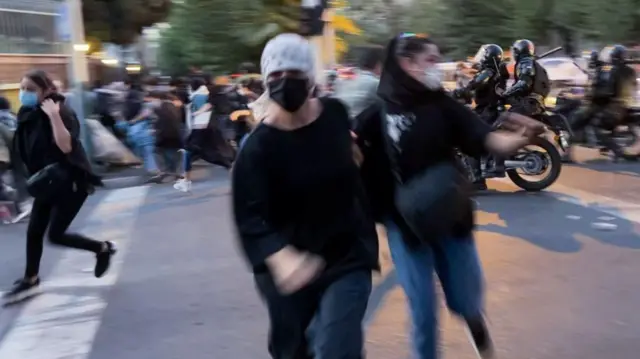 صورة ضبابية لسيدتين ترتديان كمامة، أثناء احتجاج بشوارع طهران، ويبد أنهم يركضون نحو الكاميرا، وبعض من المحتجين يهربون من شخصين يقودان دراجة نارية.