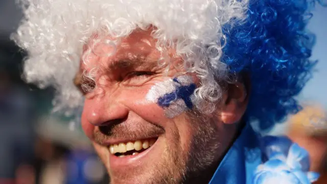 Homem sorridente ao ar livre, usando uma peruca cacheada nas cores branco e azul e com o rosto pintado com tinta azul e branca na bochecha. Ele está em um campeonato de tênis, com a luz solar direta iluminando seu rosto e outras pessoas desfocadas ao fundo.