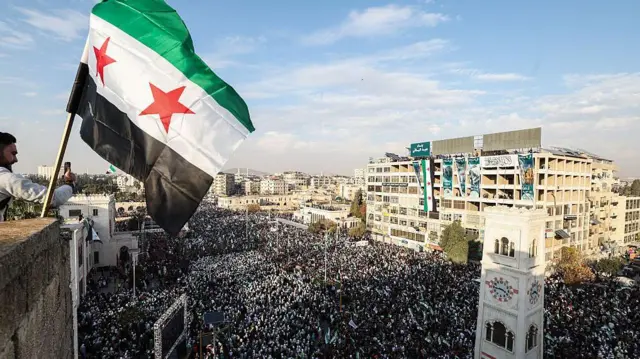 پرچم سوریه بر فراز جمعیتی در حما، در نخستین سالگرد حمله برق‌آسایی که به سقوط حاکم دیرینه کشور انجامید