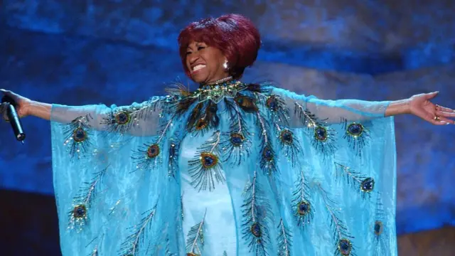 Celia Cruz, con vestido azul claro brillante, sonríe y abre los brazos mientras canta