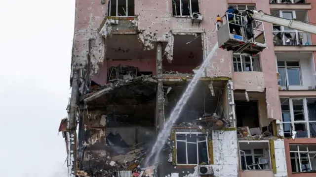 یک ساختمان دارای آپارتمان‌‌های مسکونی به رنگ گلابی با حفره‌ای بزرگ در یکی از دیوارهایش و یک آتش‌نشان که روی جرثقیل قرار دارد و به داخل آن آب می‌پاشد.