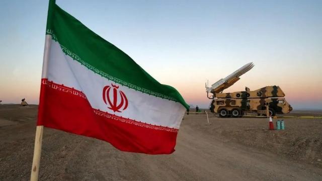 Bandeira iraniana com veículo militar ao fundo