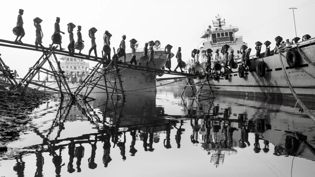 Filas de hombres transportando hacia un barco cestos llenos de carbón en sus cabezas. Las filas se ven reflejadas en el agua.