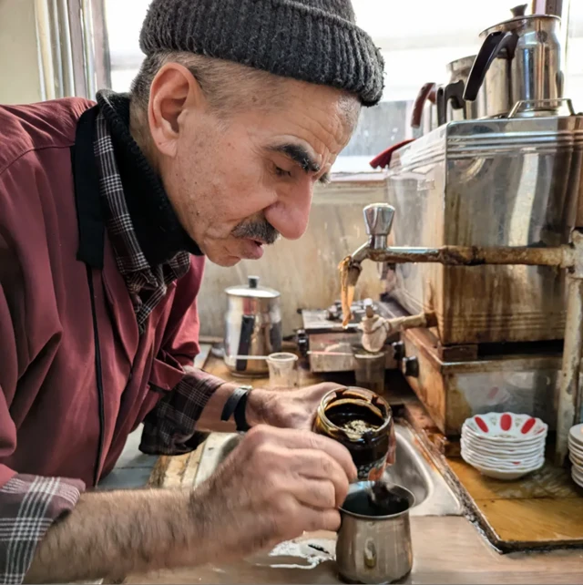 Starszy mężczyzna w czerwonobrązowej koszuli i szarej czapce wlewa gęsty, ciemnobrązowy napój do srebrnej czarki. W tle widać maszynę do parzenia kawy, ustawione na niej dzbanki i inne mniejsze naczynia dookoła.